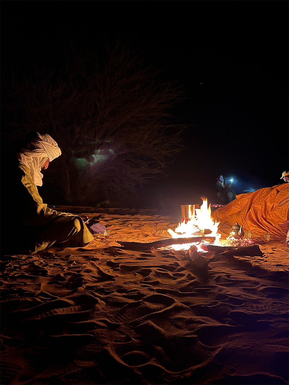 Photo: Pictured is a Tuareg making tea, a staple in their diet, in the middle of the Sahara just outside of the city of Arlit. A man dressed in tan robes form head to foot sits in front of a fire in a field of sand.