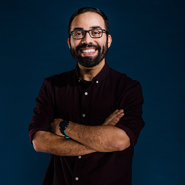 Photo: Photo of Steve Ramirez. A tan man with glasses and a beard, wearing a black collared shirt, stands in front of a dark blue background with arms crossed and smiles for the camera.