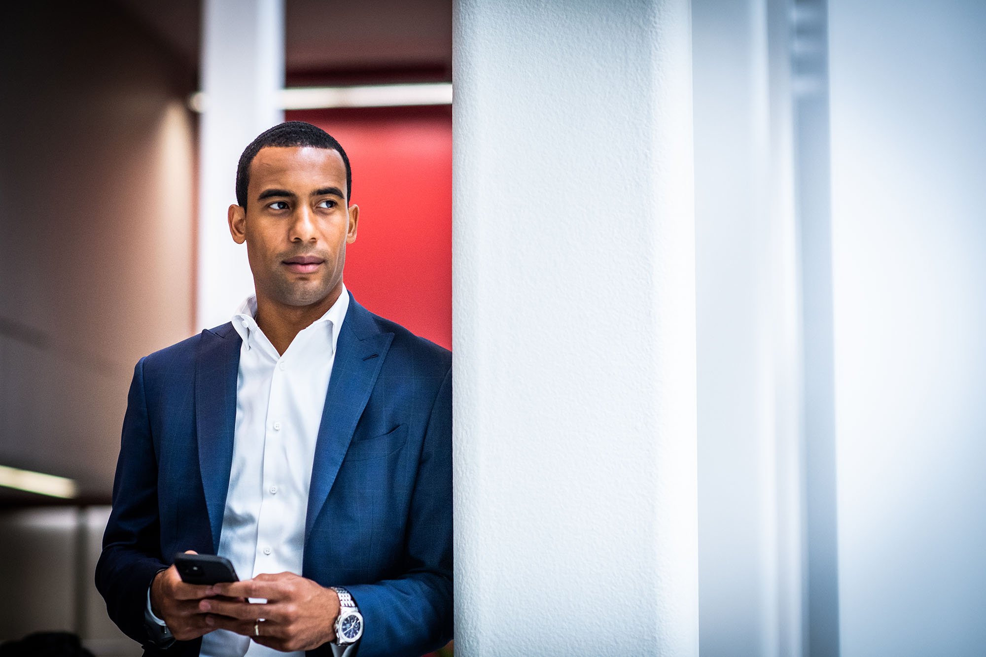 Photo of Trevor Rozier-Byrd (LAW'10) posing for a photo. A dark, tan man wearing a blue blazer and pants and white collared shirt holds his black cell phone in hand as he looks off to the right.