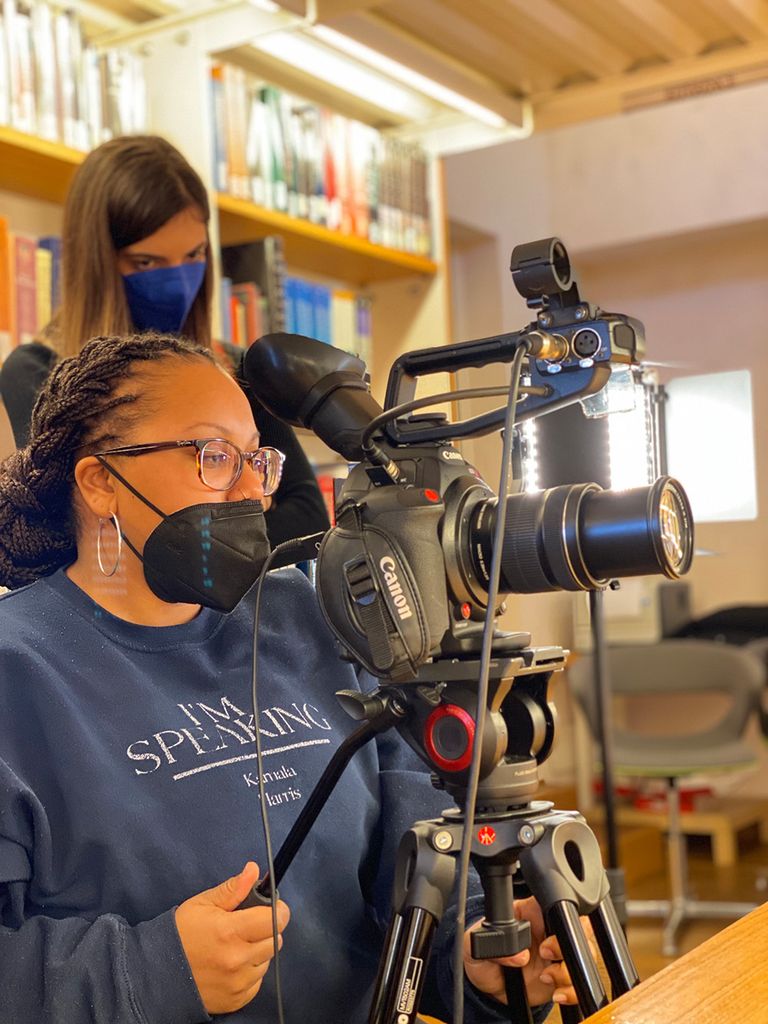 Photo of 2 young woman wearing face masks filming behind large camera setups in a library.