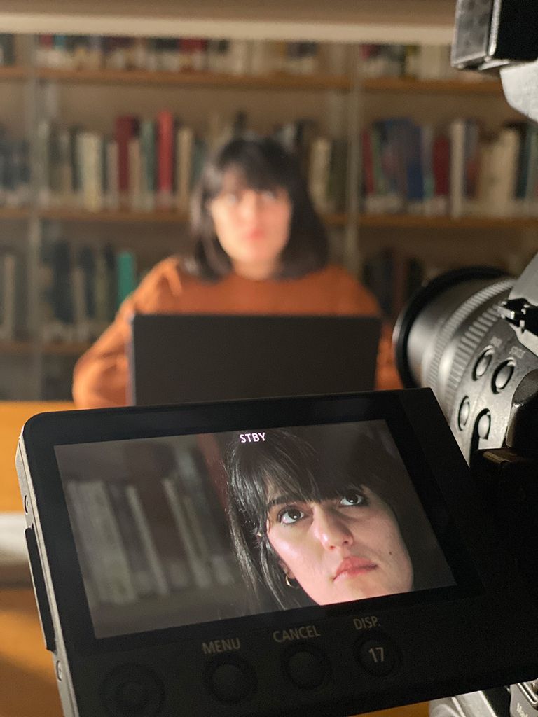 Photo of a young woman wearing an orange sweater being filmed in a library. Her face can be seen on the camera display.