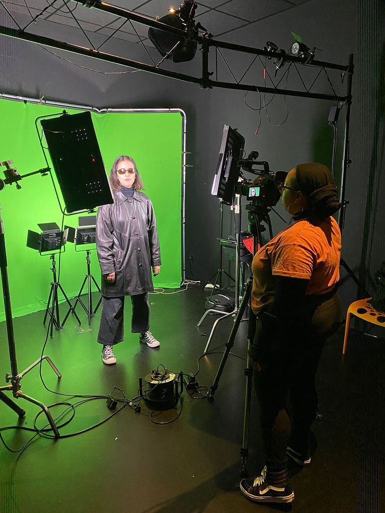 Photo of a young woman wearing all black being filmed in front a green screen in a film studio.