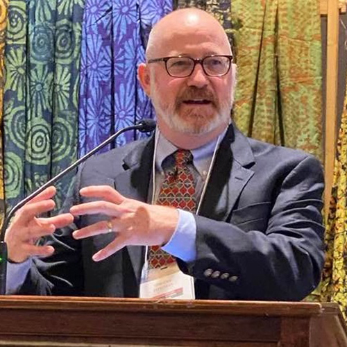 Photo: Tim Longman speaks at a podium wearing a black suit ensemble and orange tie. He gestures with his hands in front of a colorful background of drapes.