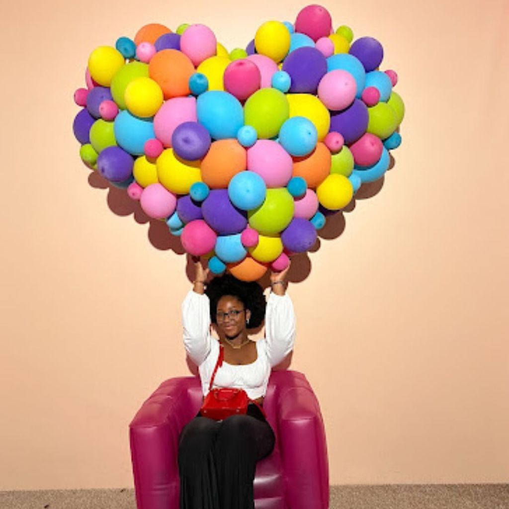 Photo of Mariame Sherazad Sow sitting in a red chair holding up a bouquet of balloons shaped like a heart.