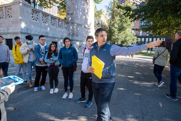Photo: Starting near the Robert Gould Shaw and the 54th Regiment Memorial, Professor Andrew Robichaud leads his History of Boston: Community and Conflict students on a tour of downtown Boston’s environmental history on October 20. A white man wearing a puffy blue vest, jeans, and long-sleeved shirt points to the right as he holds a bright yellow folder. Various students look on.