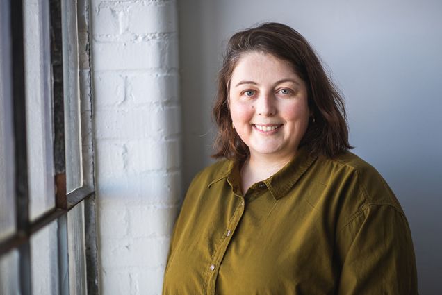 Photo: CFA professor, Josephine Halvorson poses for a photo. A white woman with shoulder length brown hair and wearing an olive green collared shirt smiles and poses next to a window.