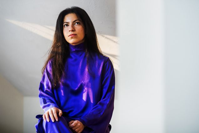 Photo: Ameera Hammouda (Questrom'18) poses for a photo at her home in Hopkinton, MA. A young woman wearing a long, dark purple dress, sits and looks up above the camera in front of a white wall.