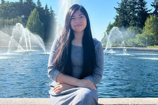 Photo: Teresa Miguel sits and poses for the camera in front of a large water fountain. A Hispanic young woman wearing a long grey maxi dress smiles for the camera.