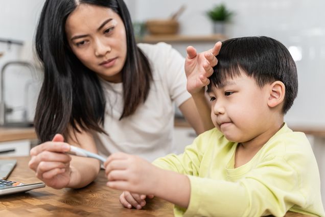 Portrait of Asian mother checking her sick sons temperature, looking at thermometer and touching forehead of her son.