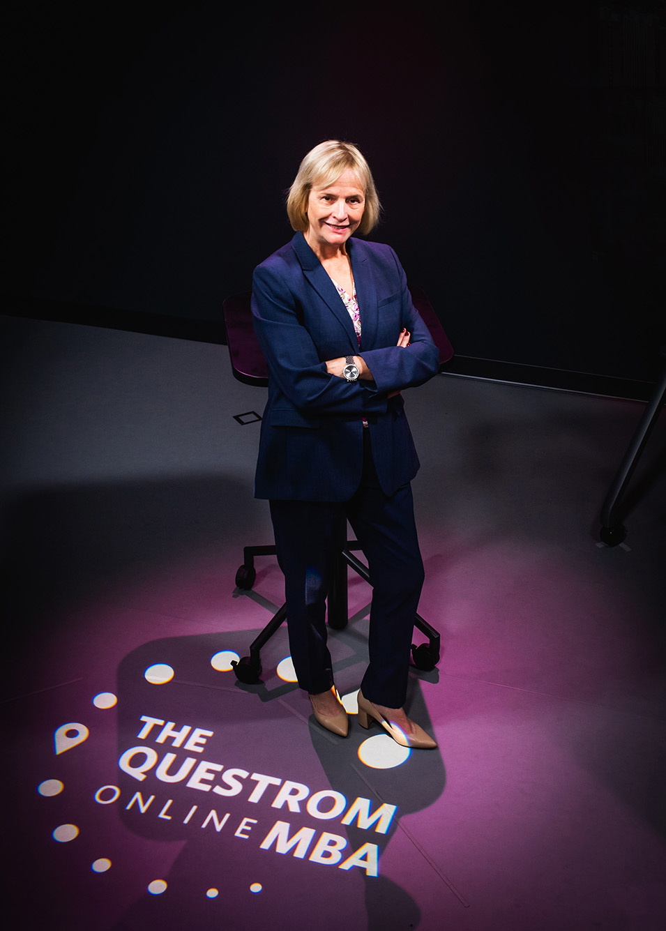 Photo: Wendy Colby, BU's first vice president and associate provost for BU Virtual, poses for a photo in a Questrom online studio. A white woman with shoulder length blonde hair and wearing a navy suit ensemble stands with arms crossed in a dark room as a purple light overlays the words "The Questrom Online MBA" at her feet.