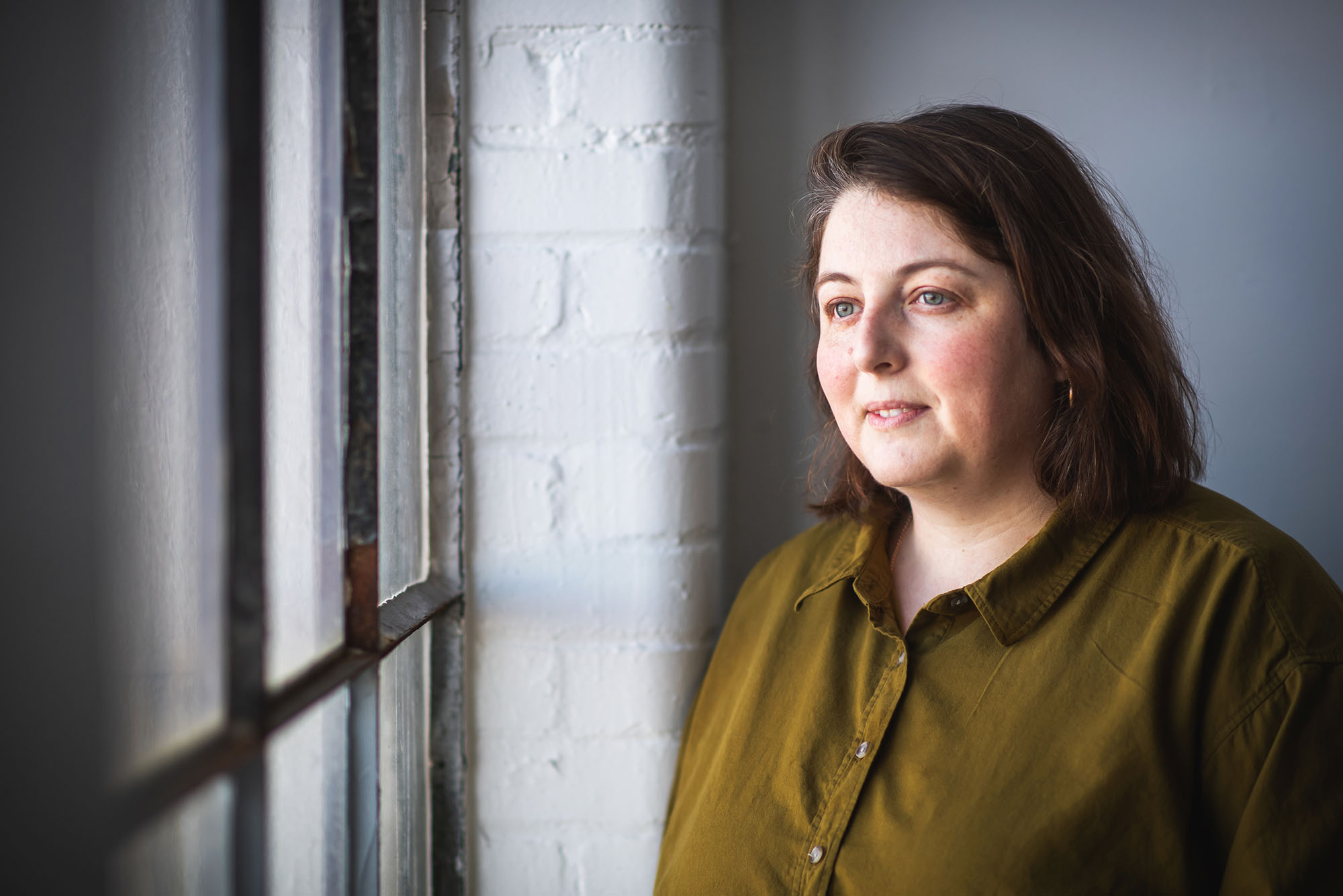 Photo: CFA professor, Josephine Halvorson poses for a photo. A white woman with shoulder length brown hair and wearing an olive green collared shirt smiles and looks out a window to her left.