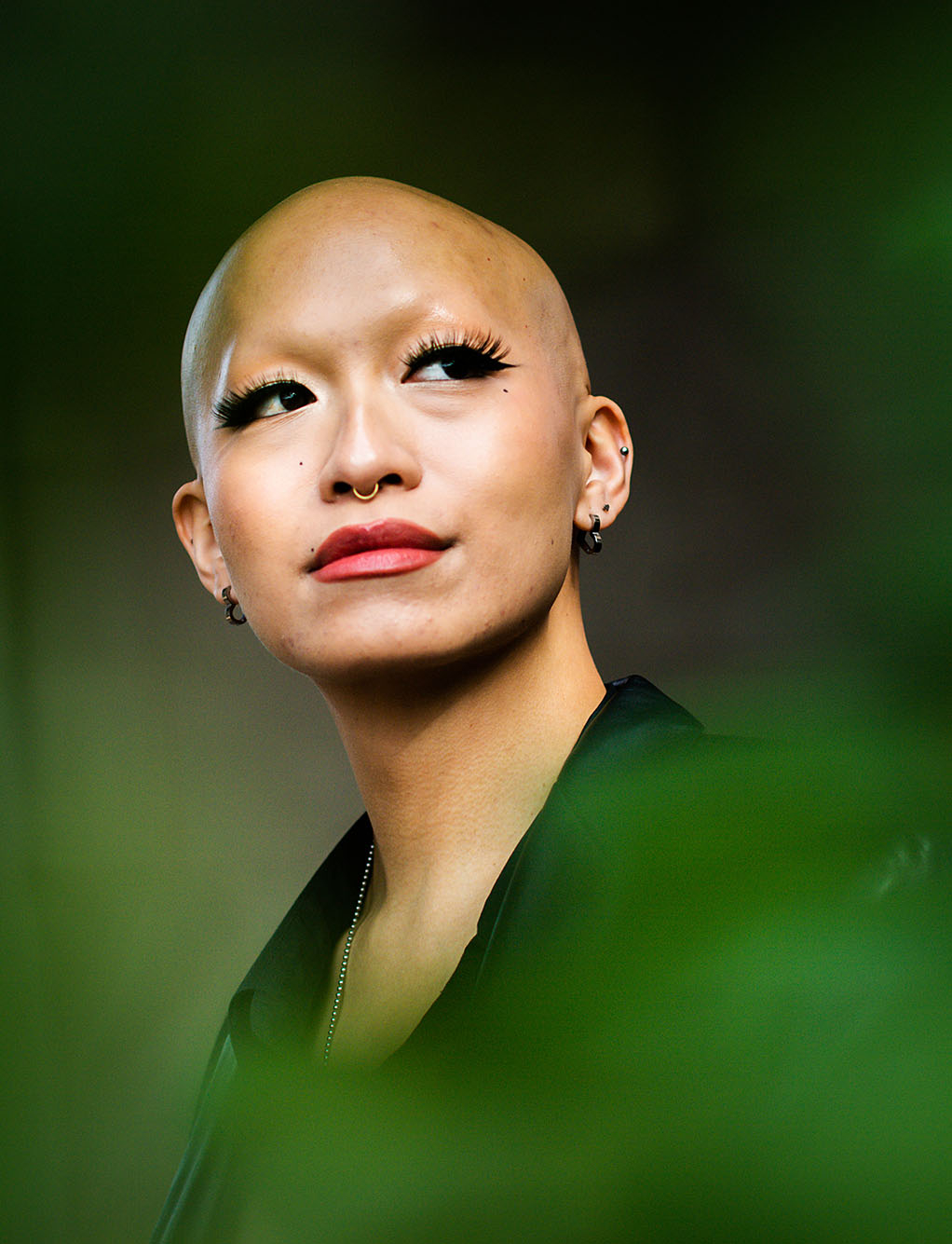 Photo: Katarina Quach poses for a photo on November 15, 2022. A young, bald, tan woman with long lashes and wearing red lipstick and a large black, oversized blazer, poses for a photo in front of a dark backdrop. Blurred, green leaves in the foreground at dimension to the photo.