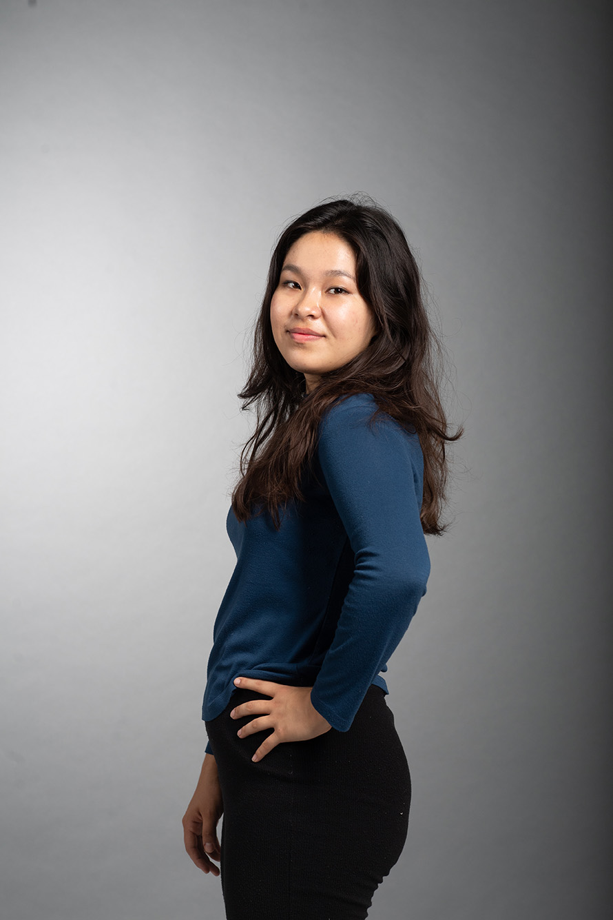 Photo: Serena Theobald poses in front of dark grey backdrop. A young woman with long black hair and wearing a navy blue long-sleeved shirt and black pants, smiles and stands sideways to the camera with hands on hips.