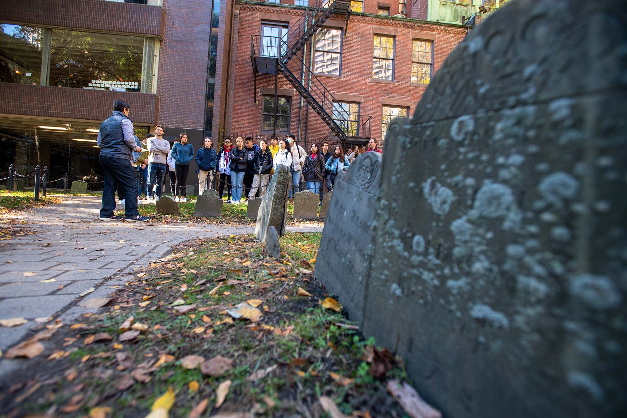 Photo: Robichaud (left) tells his students about the history of the Granary Burying Ground, where the graves of Paul Revere, Samuel Adams, and John Hancock can be found. A man faces away from the camera as he talks to a large group of students near an old gravesite.