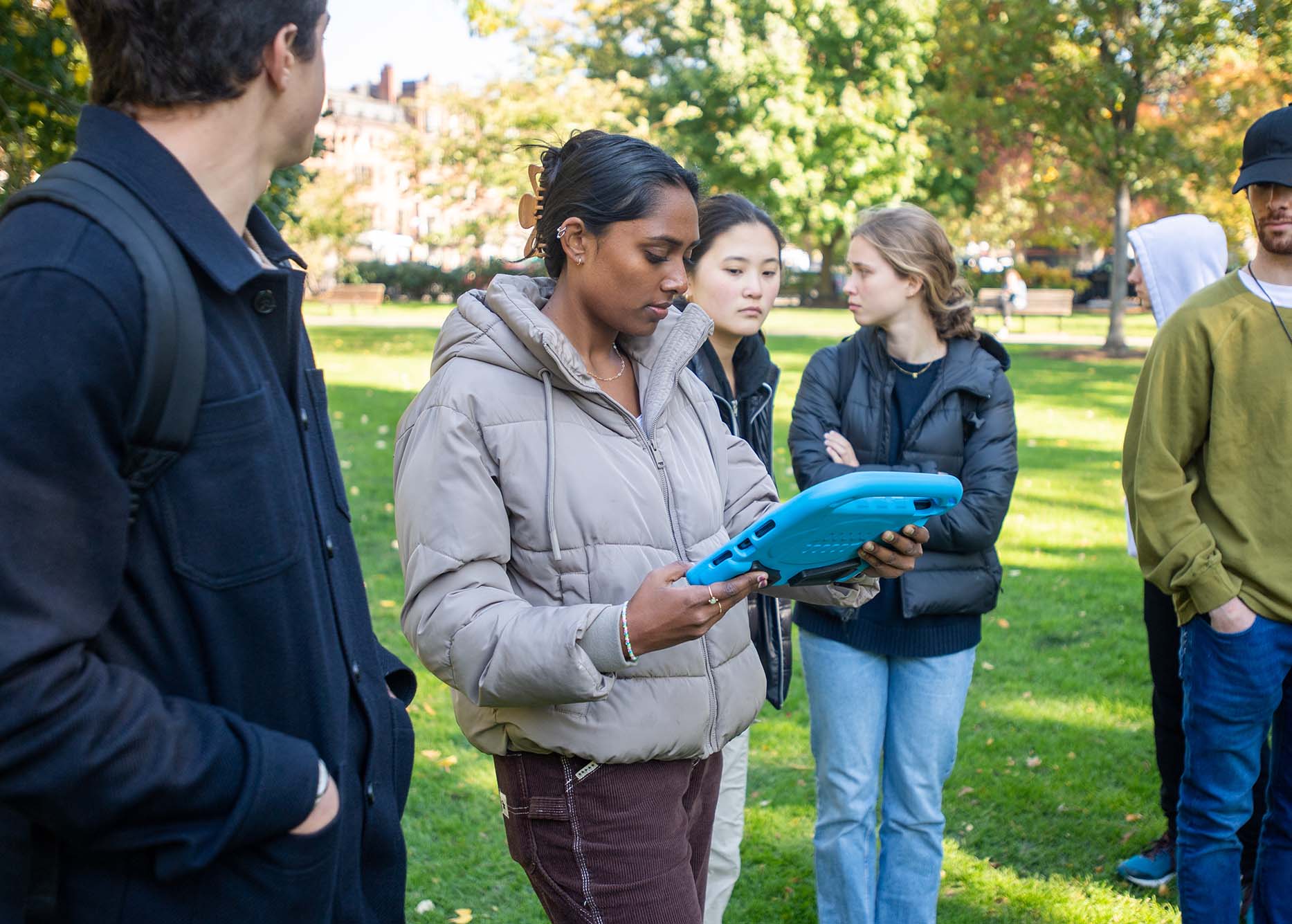 Photo: At the Public Garden, Kiran Gomatam (ENG’23) stops to look at a historical etching of the site in the 19th century. A tan young woman wearing a light lavender puffy coat looks at at etching in case by a blue protector. Various other students stand around and gaze on on the lawn