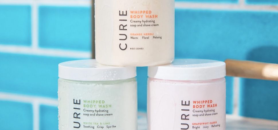 Photo: Display of three bath scrubs on a marble bathroom countertop. They are arranged in a mini pyramid with branding for the brand Marie.