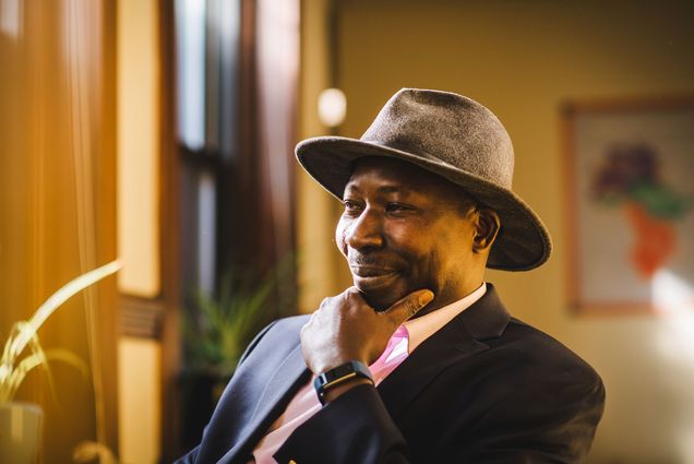 Photo: Fallou Ngom, professor of anthropology, poses for a photo on November 22, 2022. A black man wearing a grey medium-brimmed hat, light pink collared shirt, and navy blue blazer, poses with hand on chin and a soft smile as he sits in a chair.