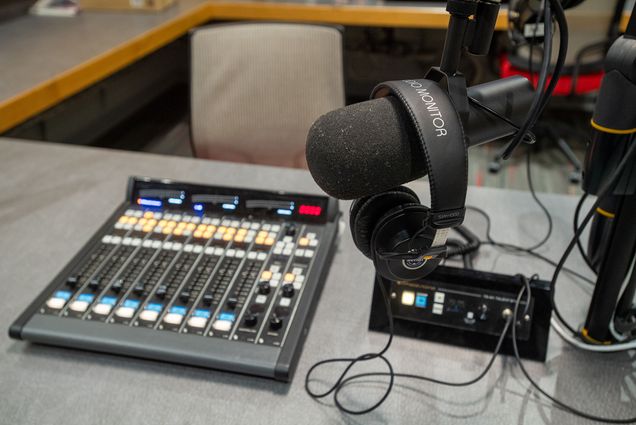 Photo: COM’s new renovated podcast studio. A grey table features a large black mixer, microphone, and professional grade headphones in a podcast studio recording room.