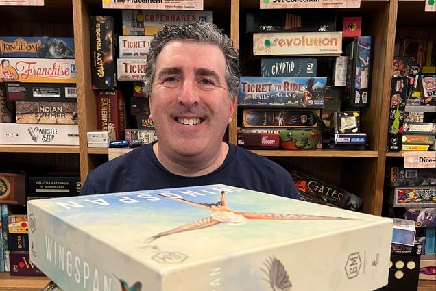 Photo: Neil Goldberg, a white man with dark silver gray hair, smiles as he sits and looks over a large stack of board games. Behind him, numerous wooden shelves filled with more board games are also shown.