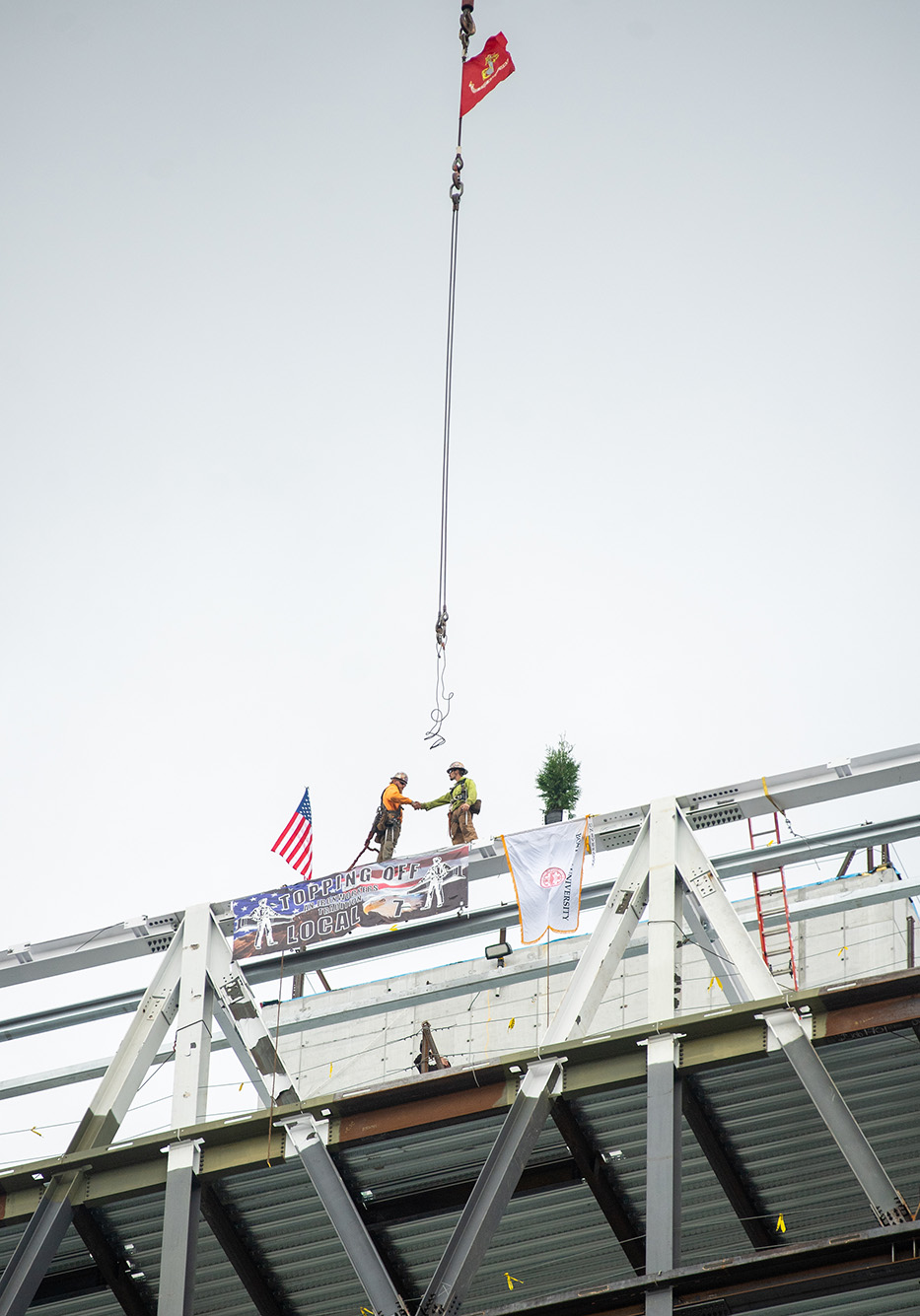 Photo: Covered in signatures, the final beam is put into place during the Topping Off ceremony at the Data Sciences Building September 30. The Marine flag and the American flag hang off the beam as two hard hat workers help lead it into place.