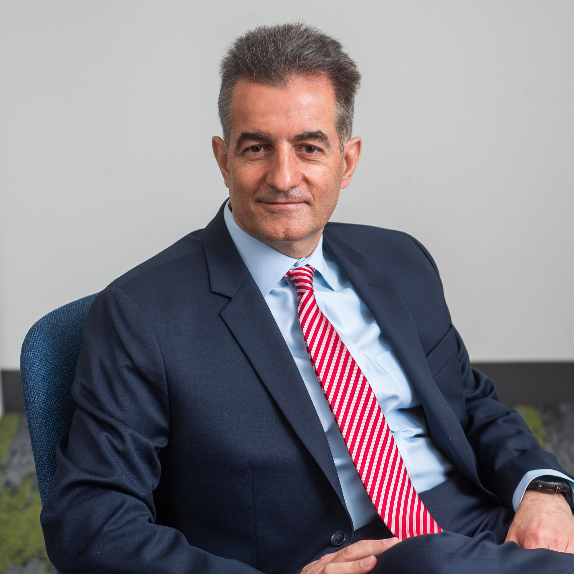Headshot of Yannis Paschalidis in a dark suit with a red tie. He sits in front of a grey background.