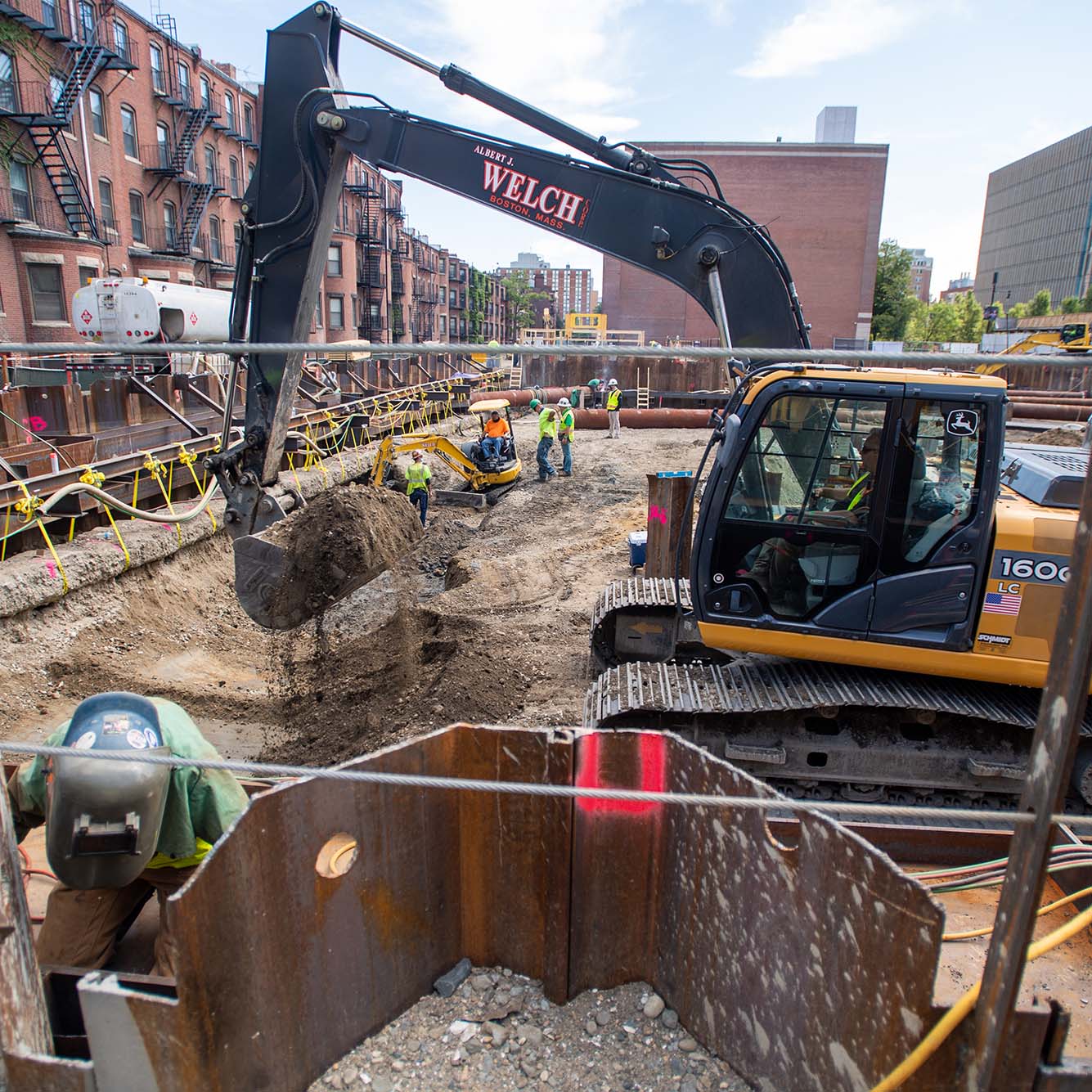 Photo: Construction site of the future Center for Computing and Data Sciences building. A large pit filled with metal work, pipes, and foundation work is shown being built in progress by construction workers.