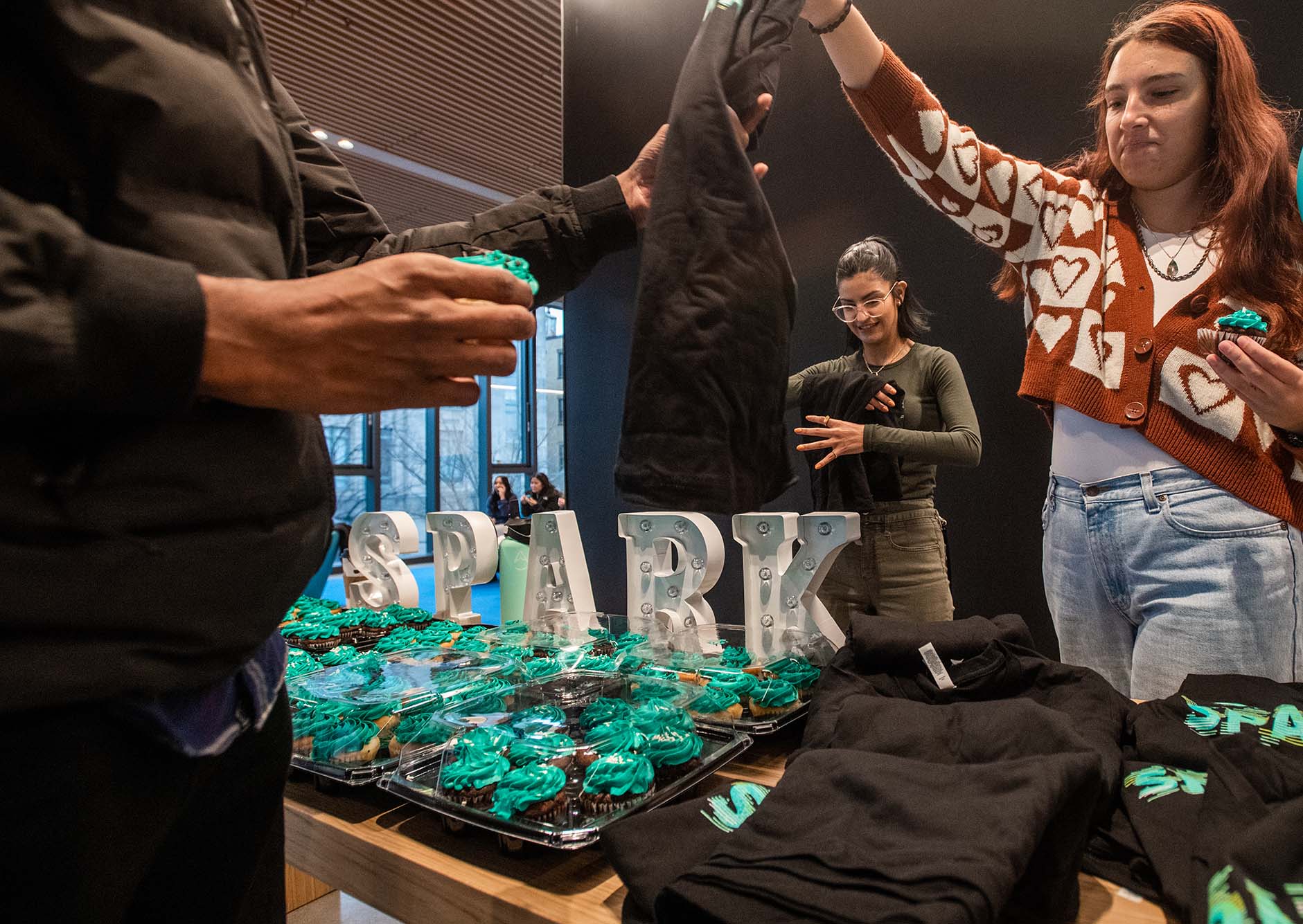 Photo: BU SPARK handed out cupcakes to visitors on opening day January 19. Two masked students hand out cupcakes decorated with bright blue frosting and black t-shirts. An unlit sign reads "SPARK" on the table.