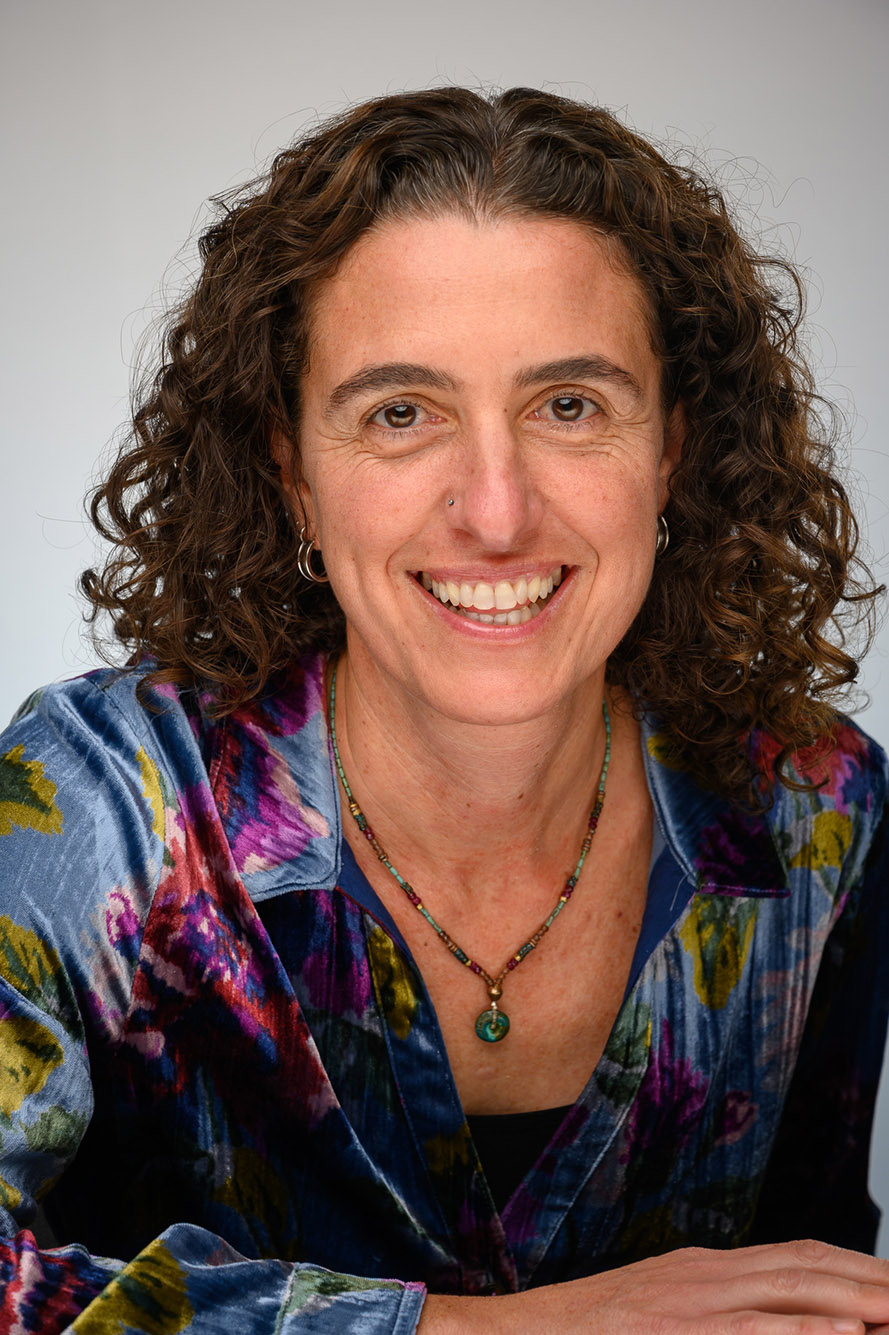 Photo": Headshot of Joanna Davidson. A white woman with curly brown hair and a colorful blue, red, and green colored shirt smiles brightly for the camera.
