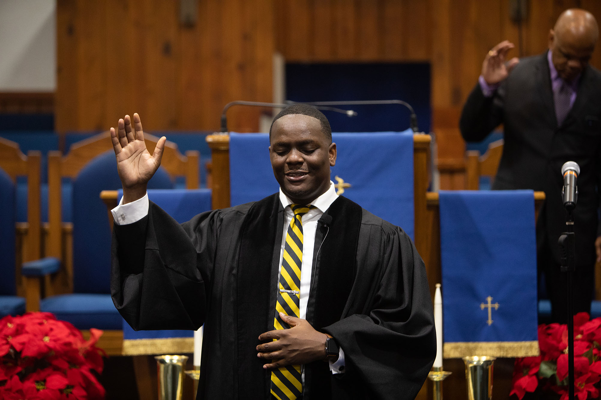 Photo: Reverend Arthur Gordon delivers his sermon at St. John Missionary Baptist Church in Roxbury. A black man wearing a black robe over a white collared shirt and yellow striped tie holds up one hand as he gives a sermon at the front of a church.