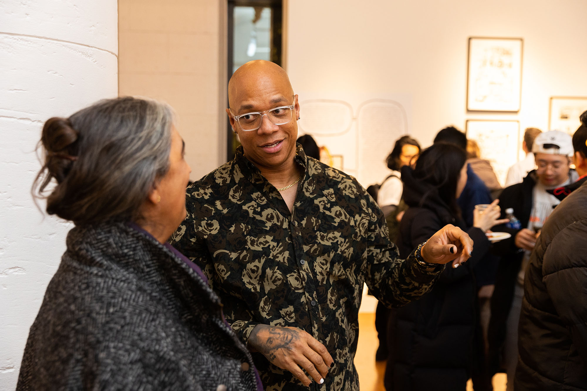 Photo: Of Joel Christian Gill, a bald Black man, in a button up shirt talking with CFA associate professor of art and director of CFA’s new MFA in Visual Narrative program. She has her hair in a claw clip. They are talking excitedly.