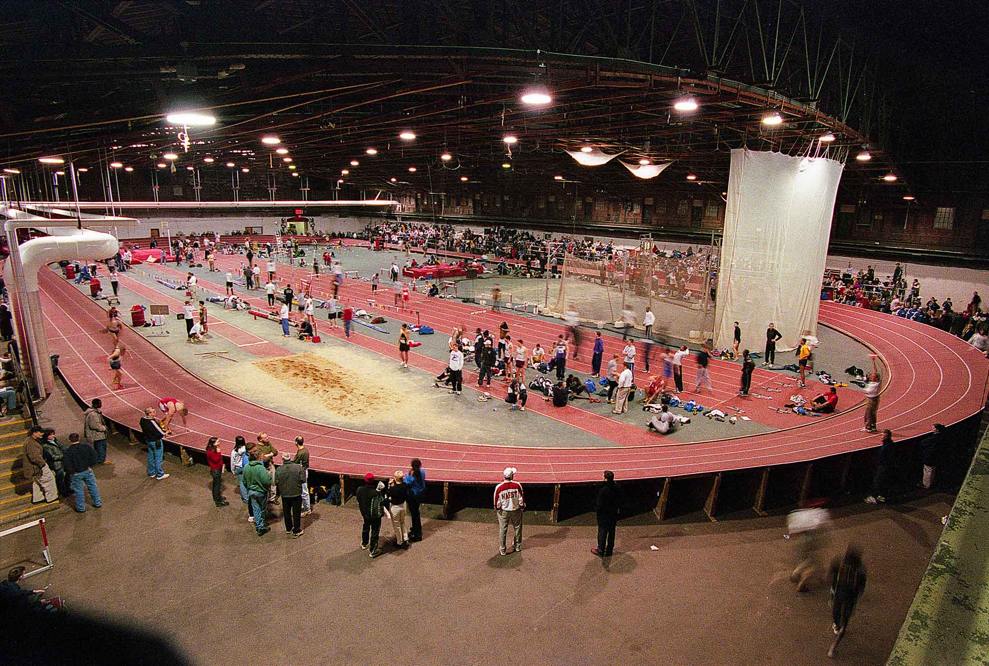 Photo: An overhead shot of a red, banked indoor track with no barriers. Scene is shown during a meet as spectators stand on the otuskirt of the track and watch as runners race on the oval. The field events can also be seen housed in the center of the track.