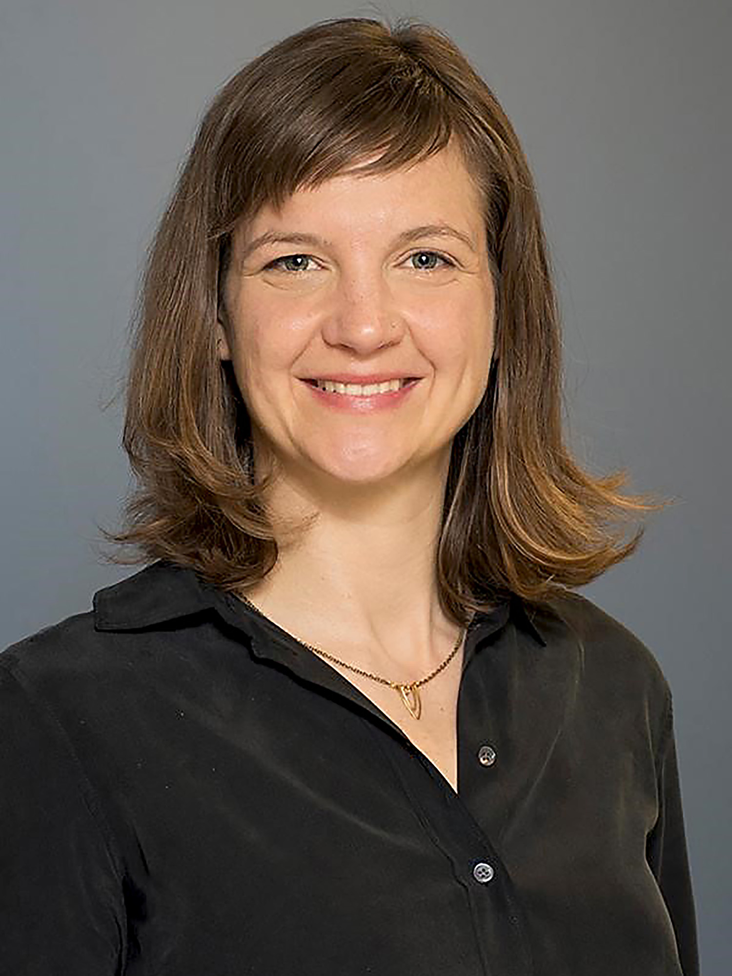 Photo: A white woman with shoulder length light brown, straight hair with bangs. She is smiling in the center of the headshot wearing a black button up blouse. She has a simply necklace on, gold, with a pendant that has two loops that mimic a "V".