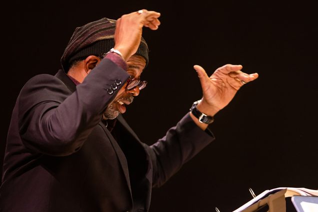Photo: Bill Banfield (STH'88) conducts a concert with his Imagine Orchestra at the Edward M. Pickman Concert Hall Longy School of Music, Friday, November 11, 2022. A Black mean wearing glasses, a purple and black knit cap, and black blazer, has his hands raised in the air as he conducts.