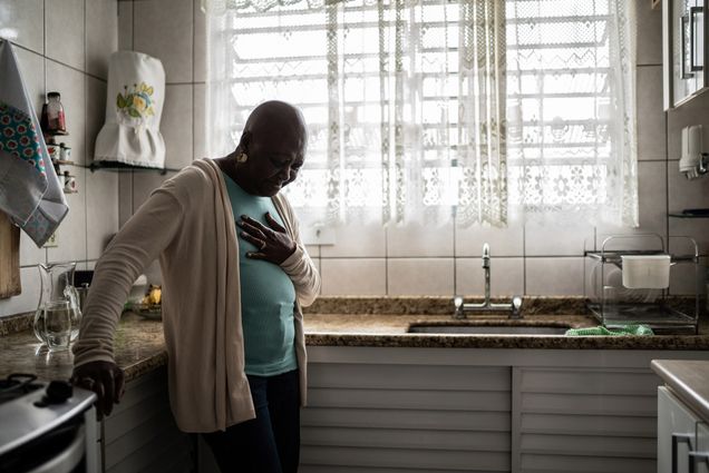 Photo: A senor, Black woman wearing a light blue shirt, tan cardigan, and black pants, has a look of pain on her face as she clutches her chest in her kitchen.