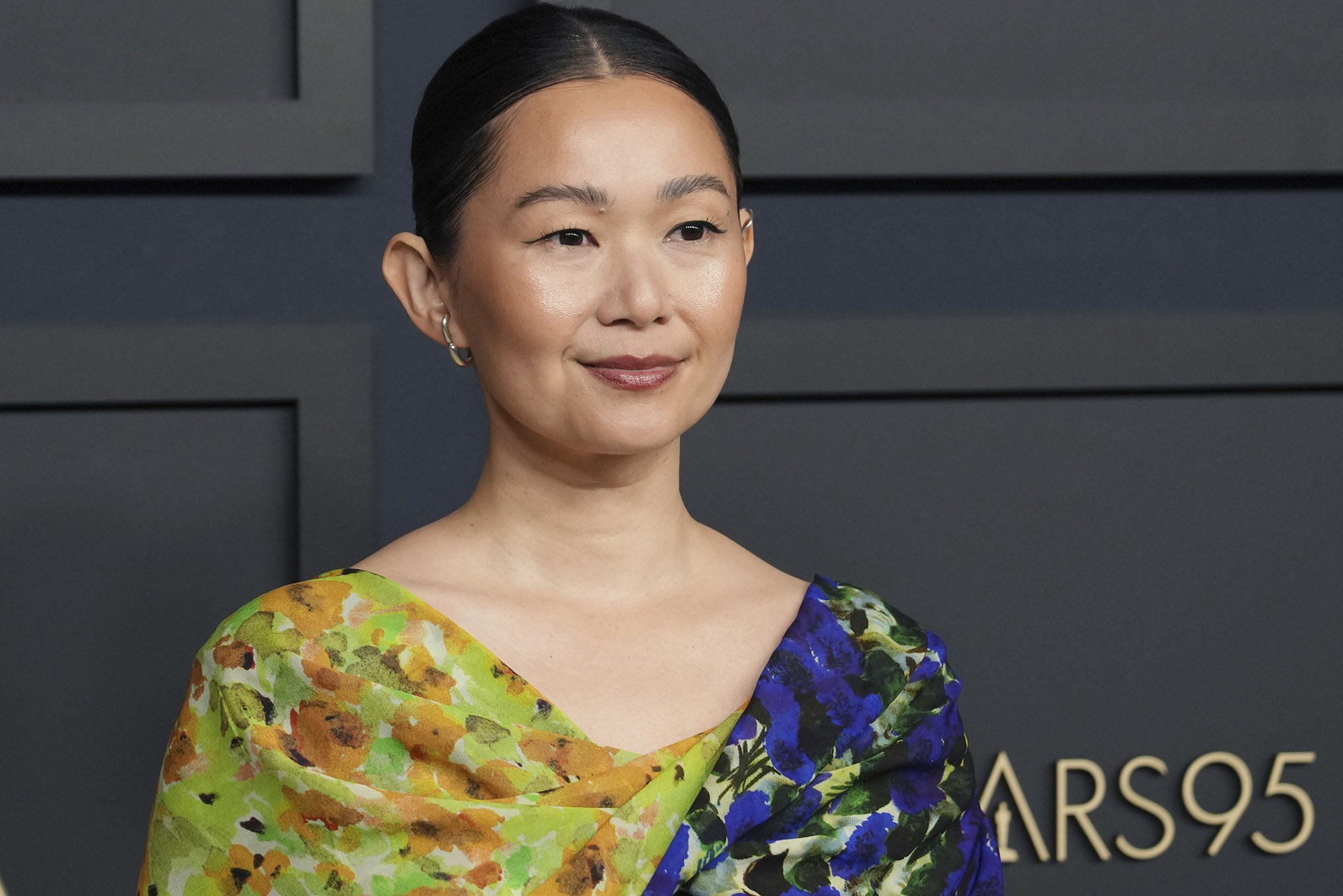 Photo: Hong Chau arrives at the 95th Annual Oscars Nominees Luncheon. An Asian woman wearing a floral, half-toned blue and green dress. She stands in front of a dark wall with a gold letter sign that reads "Oscars95"