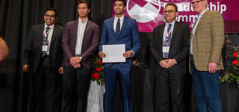 Photo: Five men stand on a stage to celebrate the winner of a compeition. From left, a West Asian man wearing a black suit, a white man in a maroon blazer and black pants, a West Asian man wearing a blue suit with a black tie, an older West Asian man wearing a black suit with a purple tie, and a white man wearing a plaid blazer with tailored jeans and glasses. They are all smiling for the photo, looking to the left at another camera.