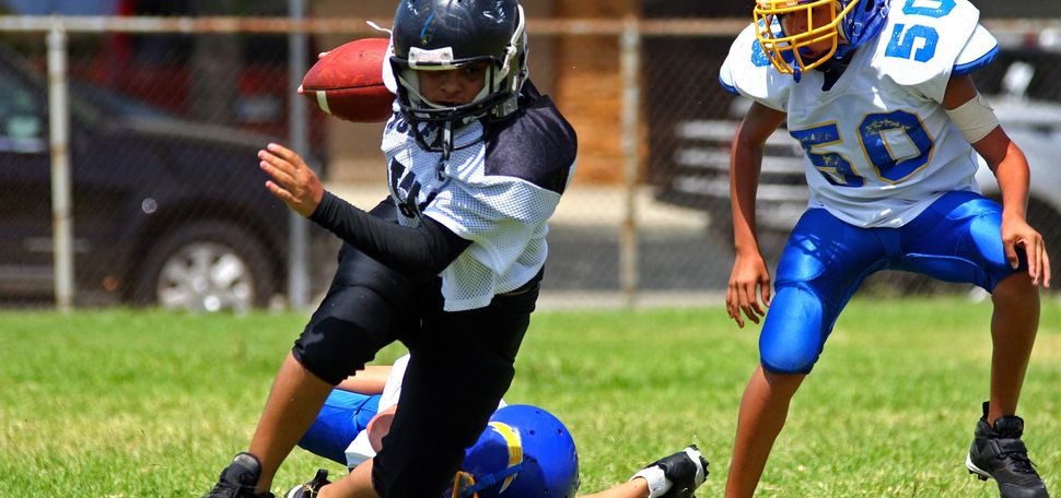 Photo: Young American football player in the running back position and wearing a black and white uniform is shown breaking away from an attempted tackle from another young player wearing a blue and white uniform. A third young player is shown in the same blue and white uniform to the right running alongside them.