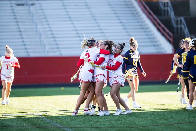 Photo: BU women lacrosse players celebrate after a goal against Merrimack in the 2023 season opener. A small group of young women wearing white and red lacrosse uniforms and sticks hug in the middle of a lacrosse field. A few young women dressed in navy and yellow lacrosse uniforms walk to their right.