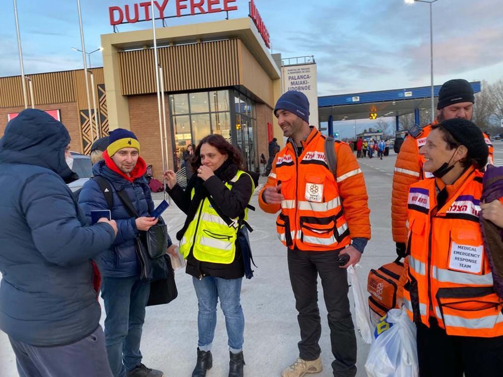 Photo of Photo of Jill Shames (COM’82 SSW’83), with a group of volunteers dressed in orange vests speaking with two men who are Ukrainian refugees. The men wear winter coats, wear backpacks and hold passports.