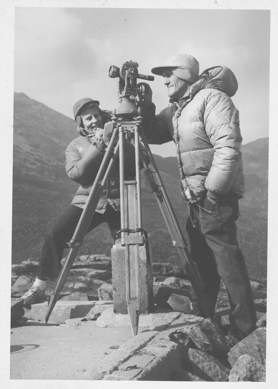 Photo: A black and white, aged photo from the Gotlieb archive of Bradford (Hon.’96) and Barbara (Hon.’96) Washburn on one of their expeditions. They both tinker with an older camera as they're dressed in bulky winter gear at the top of a mountain. 