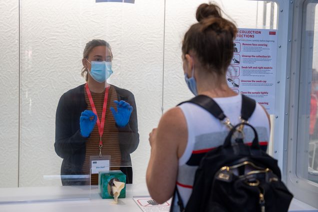 Collection site specialist Jessica Sandberg, a white woman wearing a surgical mask and blue gloves, guides a masked visitor through the COVID test collection process at the Rajen Kilachand Center.