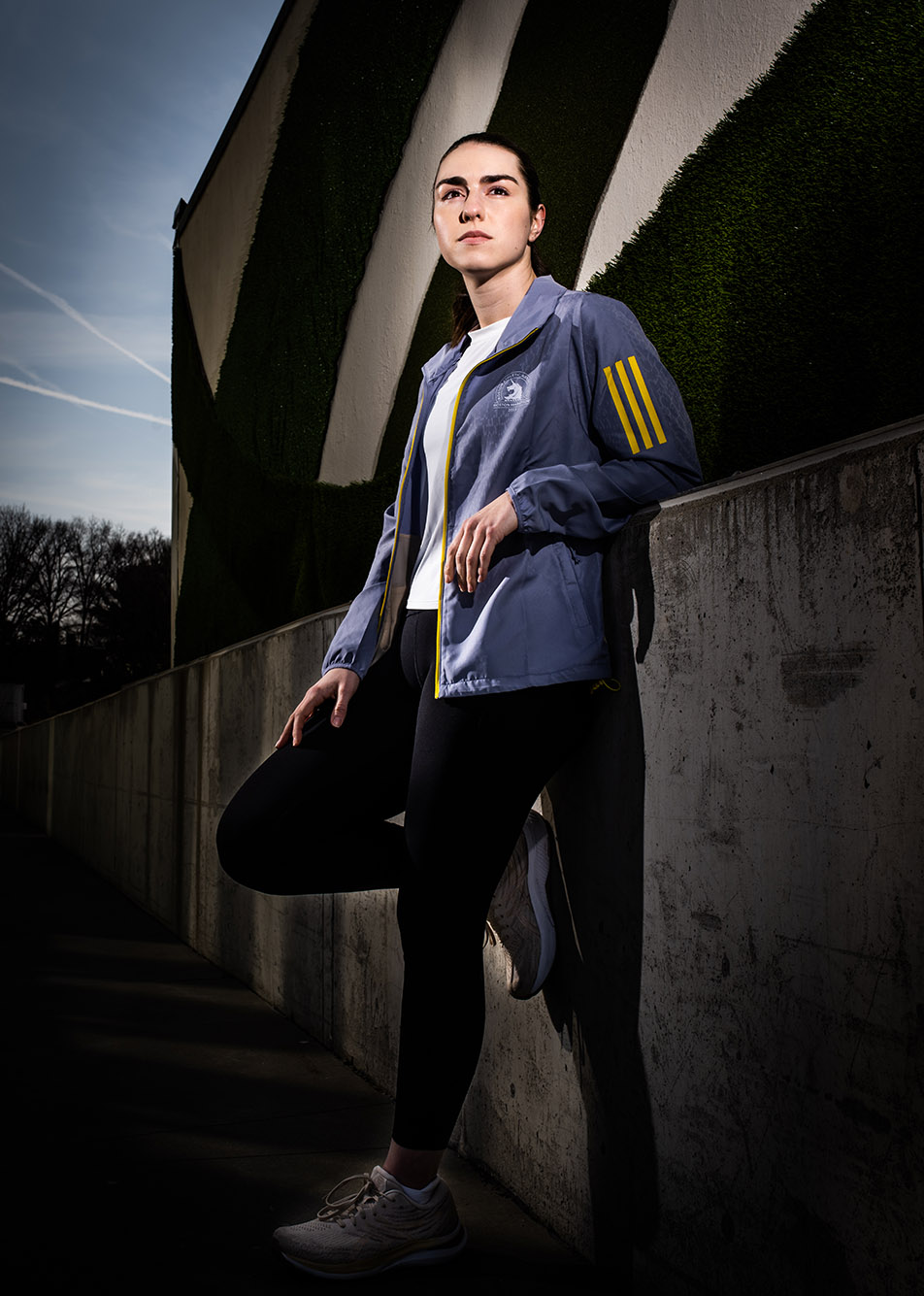 Photo: Erin Murdock poses in a moody-themed photo. She wears long black leggings and wears a light gray Boston marathon official jacket. She poses against a cement wall with one leg raised.