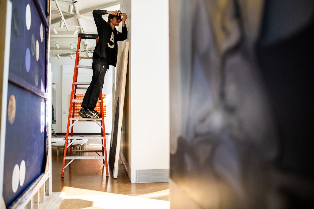 Photo: A man drills a hole into a wall to prep before hanging his canvas that lays just below. He is getting ready for the MFA thesis exhibition.