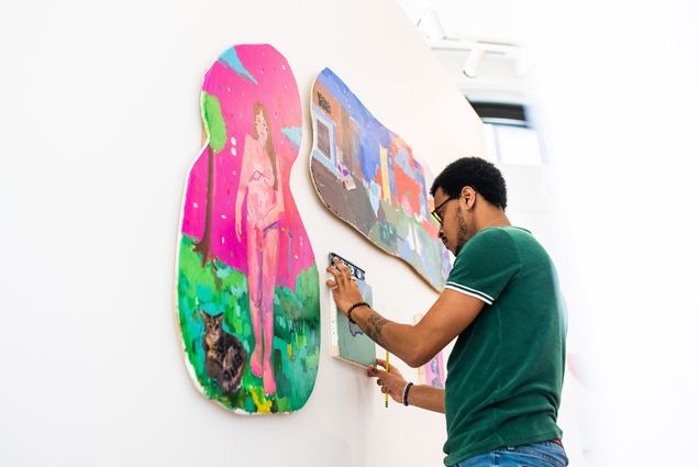 Photo: A Black man adjusts a small painting on the wall for his MFA thesis exhibition. The painting on his left is a curvy canvas (looks like an outline of the number 8) with a pink background and a nude woman on the front. The top painting is of a landscape with houses in the same form as the one on the left.