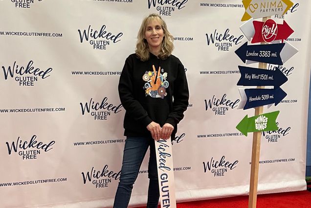 Photo: Abby Helman Kelly, a white woman with short, wavy blonder hair and wearing a black hooded sweatshirt and jeans, poses in front of a white backdrop that features a logo that reads "Wicked Gluten Free" poses with a sign in her hands that she leans on. Sign reads "Wicked Gluten Free".