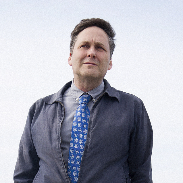 Photo: Portrait of Lecturer of Social Sciences in the College of General Studies, Sam Deese. A white man wearing a light gray collared shirt, blue polka dot tie, and dark grey jacket poses with hands in pocket in front of a large body of water.