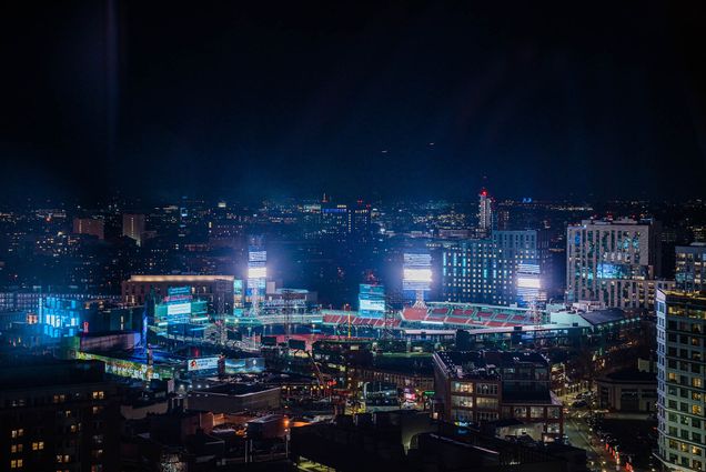Fenway Park glows with cool white stadium lights that cast a blue tint over the surrounding Back Bay Fens at night.