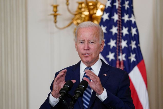 Photo: President Biden is speaking to a crowd on a mic'd podeum in a black suit with the American flag behind him.
