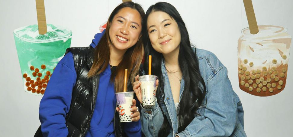 Photo: Sharlene Chang (SHA’24) and Irene Kim (COM’23), two young Asian women holding boba teas, smile and pose resting together. They sit in front of a backdrop of large boba tea recreations made out of cardboard and puff balls.