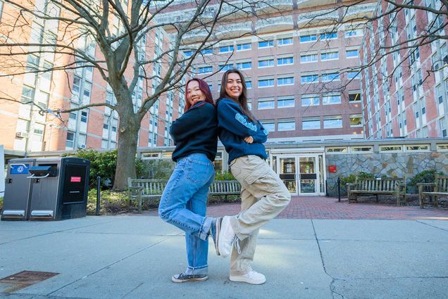 Photo: Sarah Rho (left) and Sarah Kirk poses back to back with arms crossed. Each holds their back foot against the other as the look towards the camera. The both stand in front of a large, brick, tower-style dorm residence.